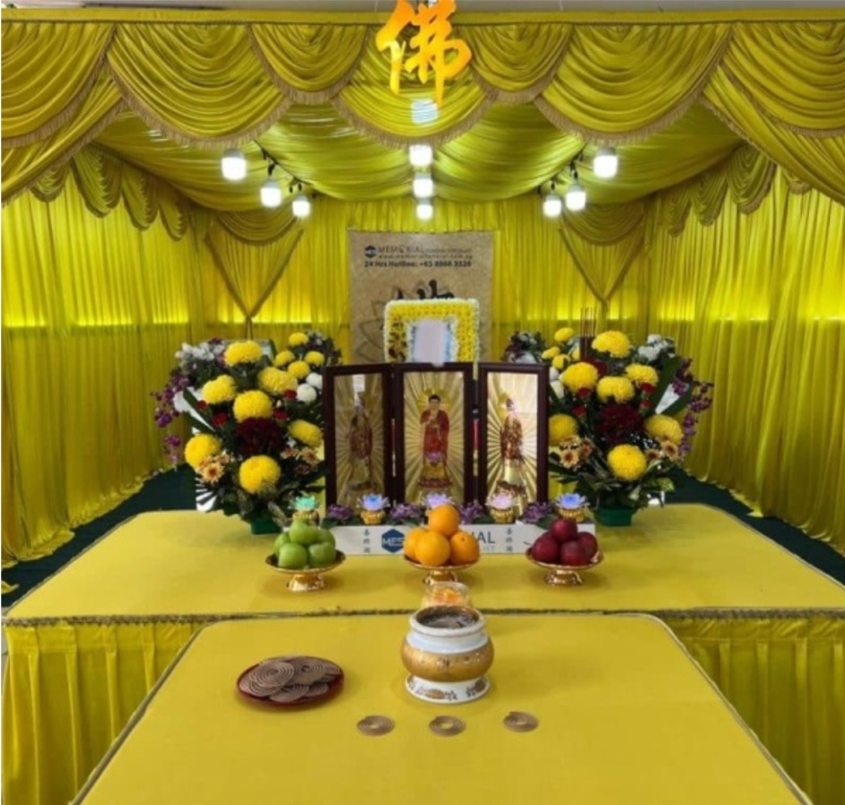 A Buddhist funeral wake setup featuring a yellow draped altar adorned with religious framed portraits, floral arrangements, offerings of fresh fruit, and an incense burner in the foreground.
