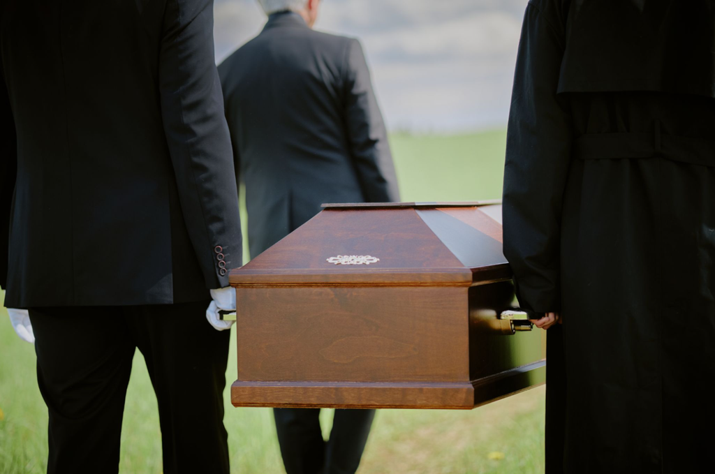 Rear view of pallbearers in black suits and coats solemnly carrying a polished wooden casket outdoors on a green lawn.