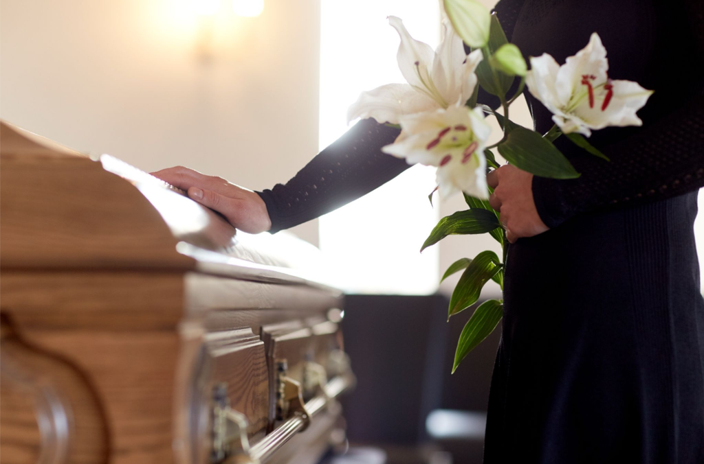 A person dressed in black gently resting a hand on a polished wooden casket while holding a bouquet of white lilies during a funeral service.